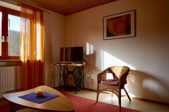 Wohnzimmer Heller Wohnraum mit einem Tisch, Stuhl, Fernseher und warmem Licht durch Fenster.